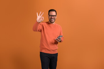 Portrait of young businessman gesturing OK sign and messaging over mobile phone on orange background