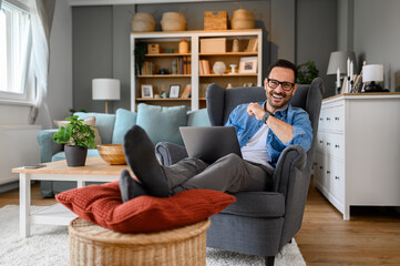 Confident male entrepreneur smiling and working online over laptop while sitting on armchair at home