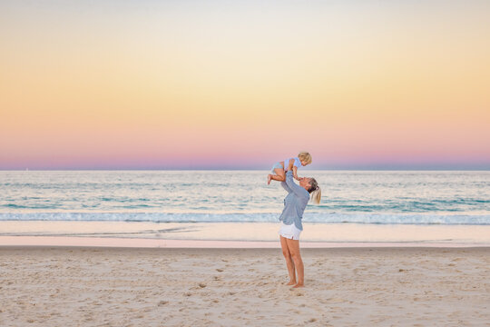 Mother lifting toddler son in the air on the beach with pink sunset sky