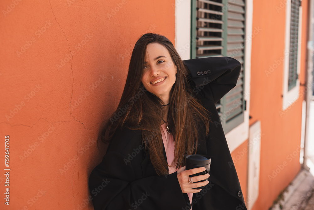 Canvas Prints Happy laughing woman standing near orange wall walking in old town, hold cup to go take away with coffee or tea. Woman drink cappuccino while rest on the street, fix her hair.