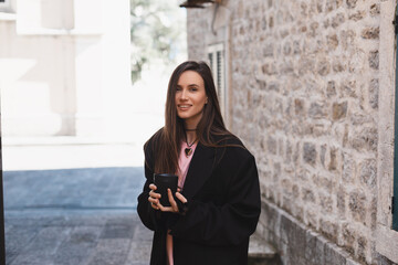 Attractive brunette woman walk on the city park street. Girl wear black jacket and look happy and smiles. Woman go on the street, she turn around in old town. Girl hold cup to go coffee