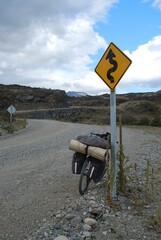 A loaded bike rests by a winding road sign amidst rocky terrain under a cloudy sky.