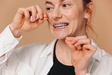 Close-up portrait of mature woman in braces holding dental floss near her teeth on beige background