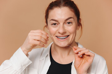 Portrait of mature woman in braces holding dental floss near her teeth on beige background
