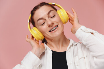 Modern woman smiling with braces on her teeth and listening to music in headphones on pink background