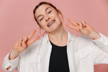 Modern happy smiling woman with braces showing peace on pink background