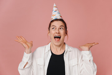 Portrait of smiling modern woman with braces and grimacing in birthday hat on pink background