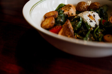A savory salad with smoked salmon, potatoes, cherry tomatoes, spinach, and a poached egg served in a white bowl.