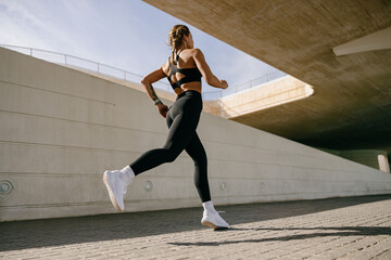 Back view of woman athlete in sportswear is running on buildings background. Active life concept - obrazy, fototapety, plakaty