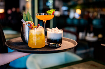 waiter holding a tray with three assorted cocktails in a cozy bar setting. The cocktails include a...