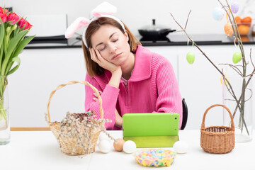 Sad woman with bunny ears headband sitting at the festive table and waiting for video call at beautiful Easter Day