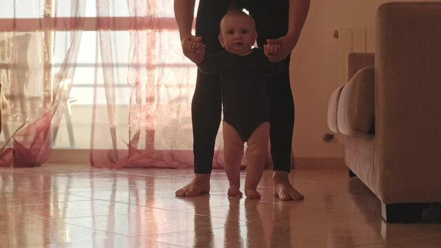 Cute baby trying to walk for the first time. Adorable infant holding his mother's hands while making his first steps. Slow-motion video of a Caucasian one-year-old boy walking for the first time.