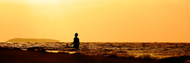 Silhouette of a young man walking in the sea (surf) towards the beach at sunset. The scene is...