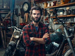 Fototapete Fahrrad Suggestive portrait of a young handsome mechanic in a red-checked shirt standing in his vintage authentic bike shop among motorcycles.