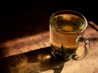 A transparent cup of tea with green tea stands on the table and is illuminated by the sun