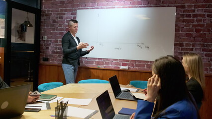 Office workers listen intently to brunette man at whiteboard. Young manager gestures explaining graph on whiteboard to colleagues