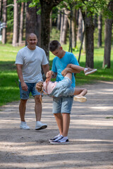 Fototapeta premium A boy playfully upends a giggling younger child as an adult looks on, sharing joy in a sunny, tree-lined park, National siblings day and friendships.