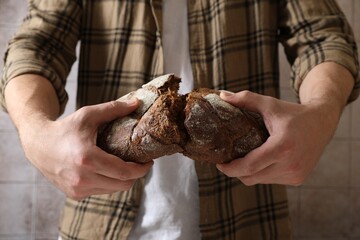 Man breaking loaf of fresh bread near grey wall, closeup