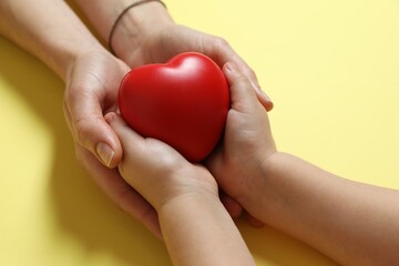 Mother and her child holding red decorative heart on pale yellow background, closeup