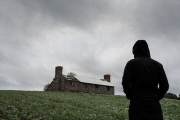 A mysterious hooded figure looking at an abandoned ruined farm house on a hill. On a grey stormy...