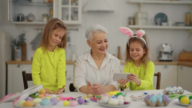 Easter grandmothers with granddaughters. Smiling grandmother with twins grandchildren watching cartoons, paint lesson on phone in rabbit ears, talking, celebrating together at home. Easter concept.
