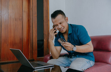 Happy Indonesian man buying things online, using smartphone, laptop and credit card, enjoying shopping in internet. Positive young guy purchasing goods on web, making remote payment.