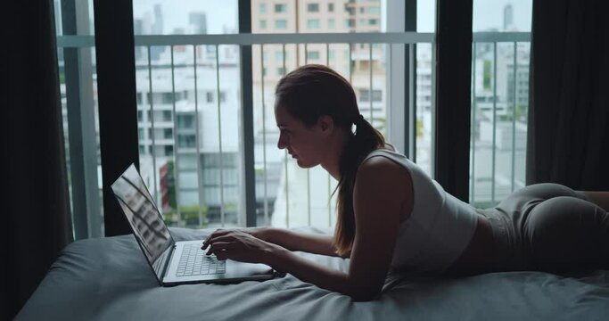 Woman works on laptop lying on bed in bedroom close-up side view Work from home merging living space with office. Work from home online revolution Work from home flexibility in apartment.