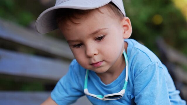 Charming baby boy in cap, blue t-shirt and with sunglasses is on the bench outdoors. Cheerful kid stands up and walks away. Close up. Blurred backdrop.