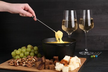 Woman dipping piece of bread into fondue pot with melted cheese at black wooden table, closeup