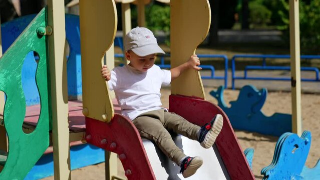 Energetic baby sitting on the slide on sunny day. Kid in cap bangs his feet by the slide on the playground.