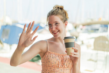 French girl with glasses holding a take away coffee at outdoors saluting with hand with happy...
