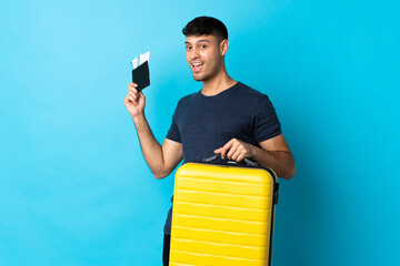 Young Colombian man isolated on blue background in vacation with suitcase and passport