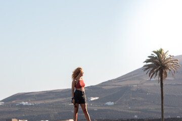 A woman stands on a hill overlooking a palm tree. She is enjoying her vacations on lanzarote. The...