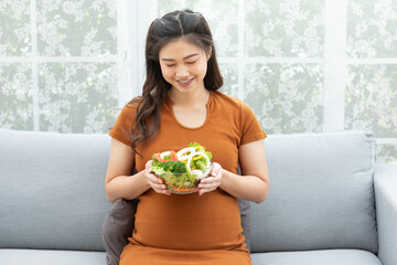 Smiling pregnant woman holding a bowl of fresh vegetable salad, enjoying a nutritious meal at home