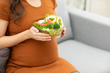 Smiling pregnant woman holding a bowl of fresh vegetable salad, enjoying a nutritious meal at home
