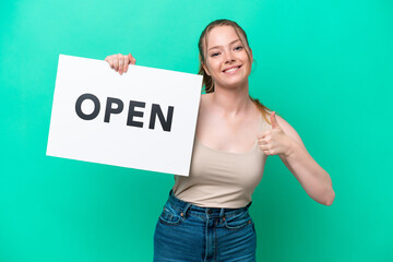 Young caucasian woman isolated on green background holding a placard with text OPEN with thumb up