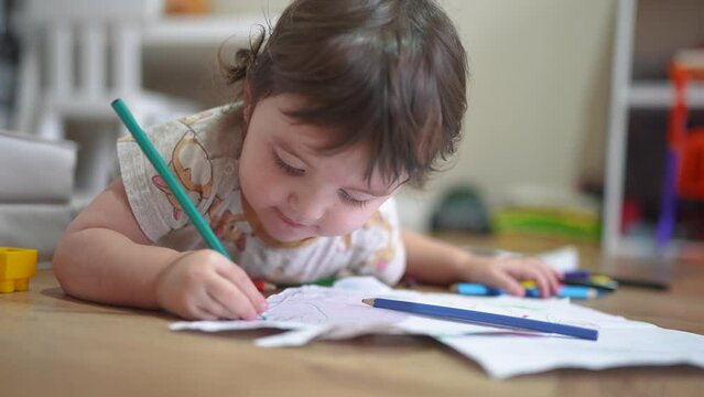 child draws with pencils on a sheet. happy family child dream concept. baby girl lies on the floor indoors and draws pictures , developing fine motor skills. lifestyle of drawing a child