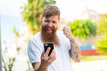Redhead man with beard using mobile phone and doing victory gesture