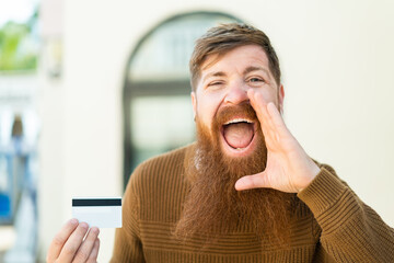Redhead man with beard holding a credit card at outdoors shouting with mouth wide open