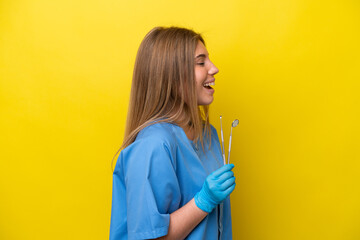 Dentist caucasian woman holding tools isolated on yellow background laughing in lateral position
