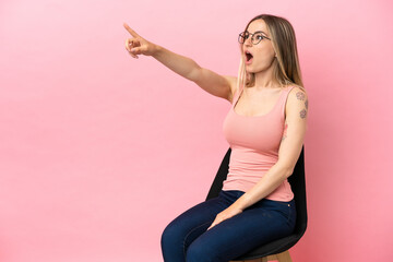 Young woman sitting on a chair over isolated pink background pointing away