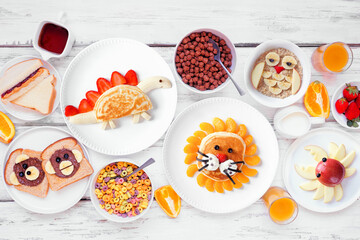 Fun child theme breakfast table scene with a selection of animal themed foods. Above view on a white wood background. Pancakes, oatmeal, toast, fruit and cereal.