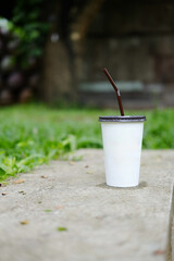 Paper glass of iced coffee on concrete step floor with grass field in tropical garden of cafe