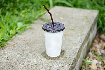 Paper glass of iced coffee on concrete step floor with grass field in tropical garden of cafe