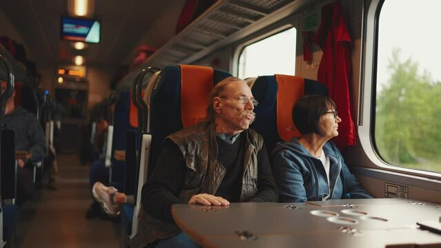 Elderly married couple in glasses look out the window while traveling by train, Slow motion. An old husband and wife look out the window while riding on a train.