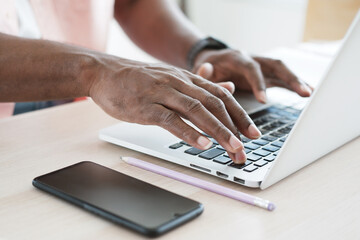 Closed up on hands of African businessman is working and typing on laptop keyboard for online meeting on wooden desk in modern office. Business and technology with smartphone concept