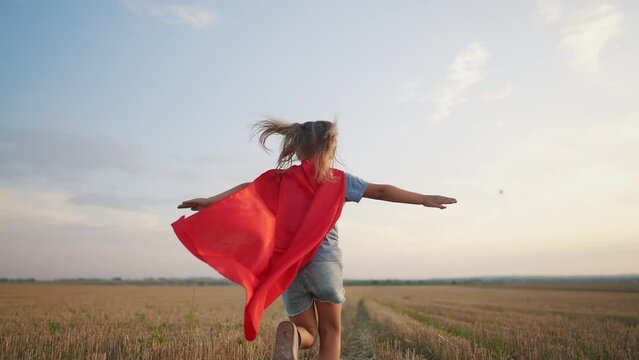 girl with the cloak. concept of a happy childhood and family for a child. a little girl in a superhero costume runs across a field, a red cape and a blue T-shirt, sunset on the lifestyle background