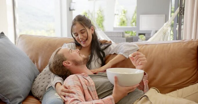 A young biracial couple shares a moment of laughter on a sofa at home, eating popcorn