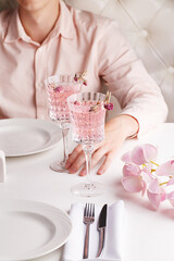 Restaurant table served with tableware, orchid flowers and two glasses of pink cocktail decorated with rose buds. Man sits at the table and holds a glass in his hand