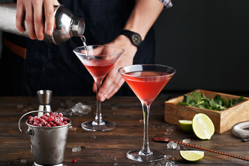 Bartender pouring Cosmopolitan cocktail in martini glass. Two glasses of red cocktail with ingredients on wooden table. Side view